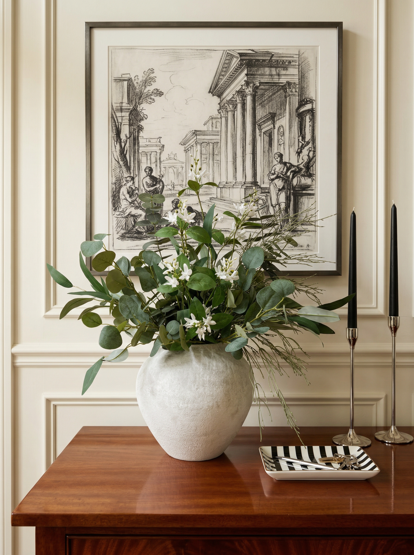 Decorative arrangement of greenery in a vase on a wooden table with framed artwork in the background.